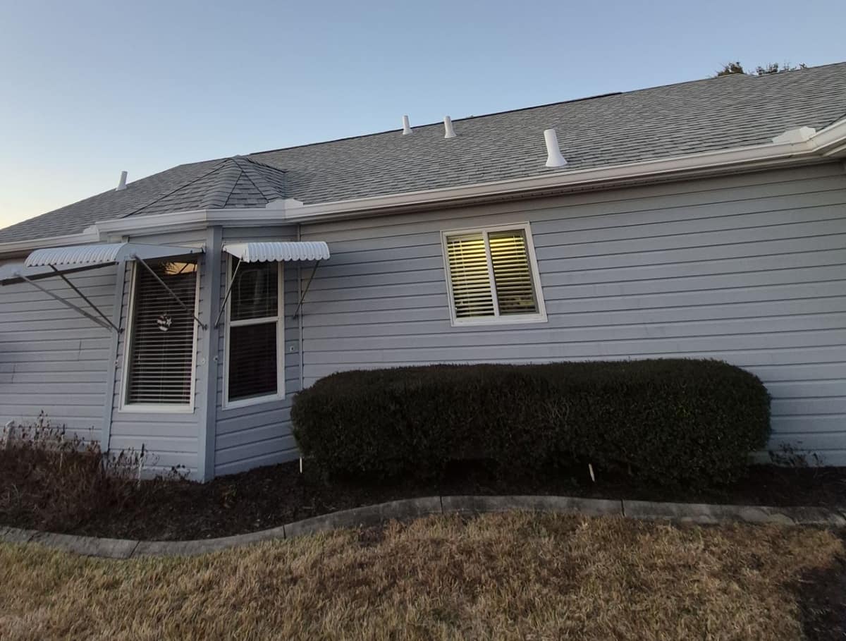 Gray-sided Dunnellon home with window awnings, a trimmed hedge and white pipe vents on the new roof