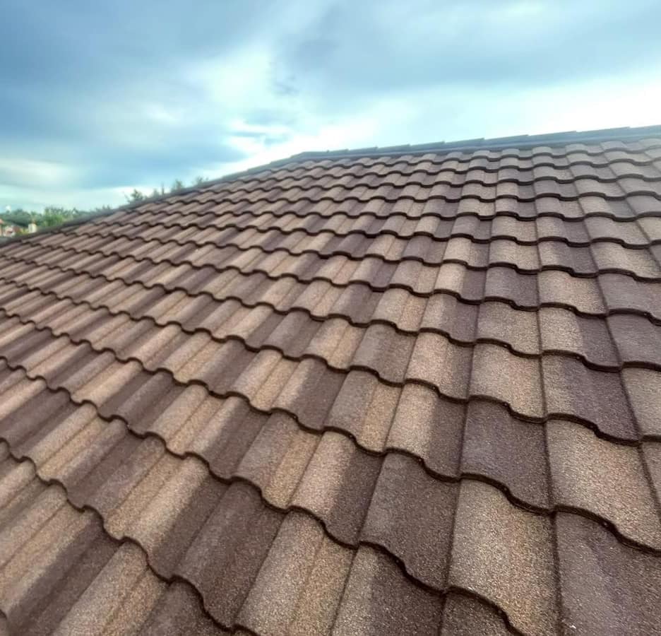 Wide slope of barrel-shaped metal tile under a cloudy sky, with neighboring roofs beyond