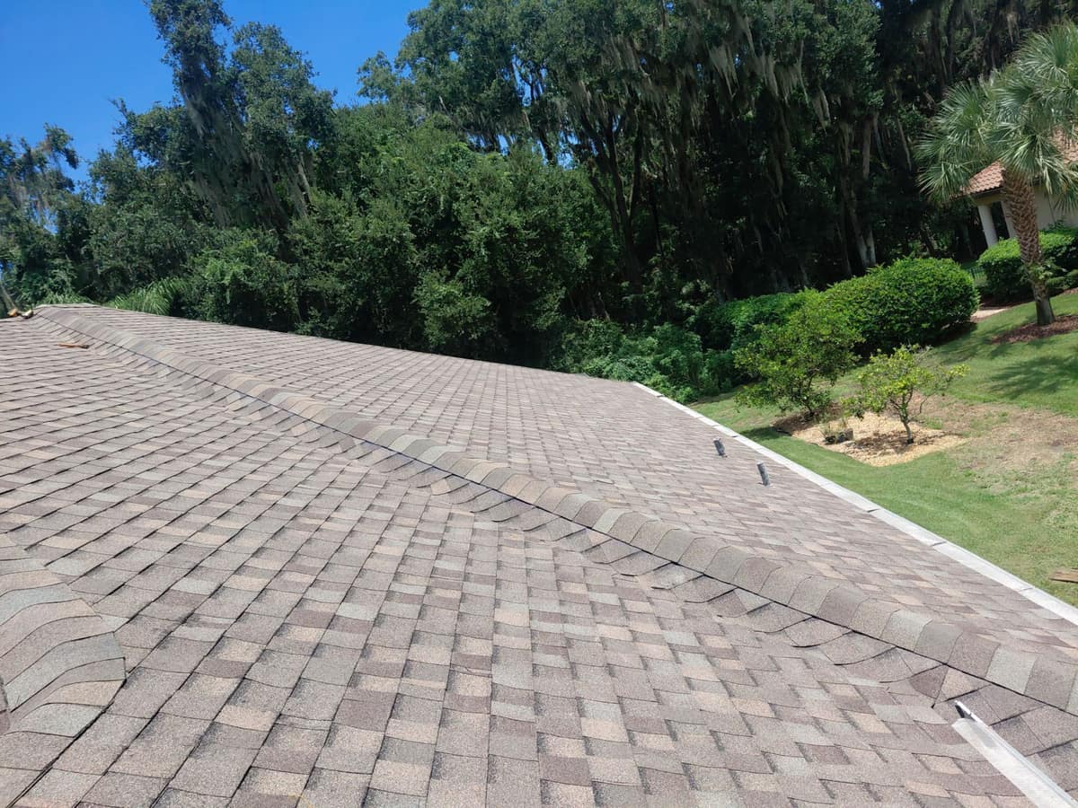 Ridge caps and two black pipe vents on the brown blend roof, mossy oaks past the lawn